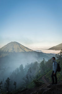 Man standing on cliff with volcano in background against blue sky