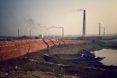 Smoke emitting from stack in front of lake against sky on sunny day