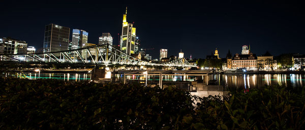 Illuminated modern buildings by river against sky at night