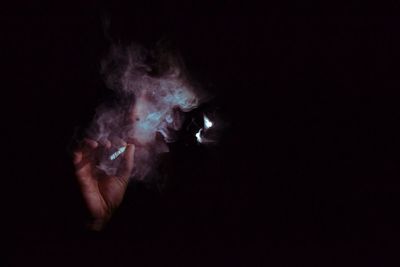 Close-up of person holding cigarette amidst smoke against black background