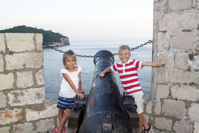 Smiling siblings standing by cannon against sea