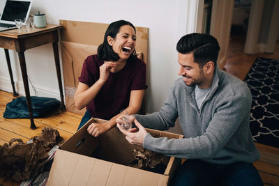 Laughing couple unpacking cardboard box while sitting on floor at home
