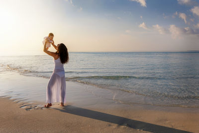 Mother playing with daughter while standing on shore at beach against sky during sunset