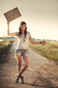 Full length portrait of woman hitchhiking while holding text on road