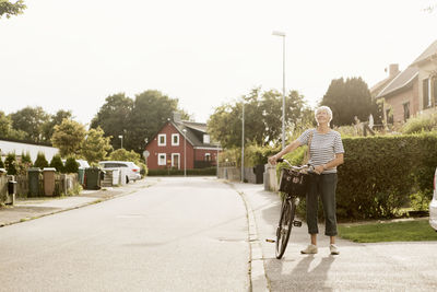 Full length of senior woman with bicycle standing on sidewalk by houses