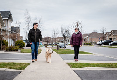 Man and older lady walking dog on sidewalk of suburban neighbourhood.