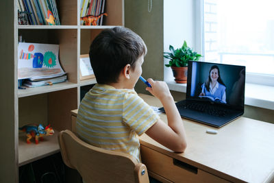 7 years old boy sitting by desk with laptop doing writing task during online lesson. side view