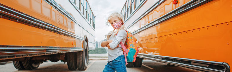 Girl kid student in face mask standing by yellow school bus. new normal. web banner header.