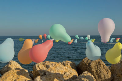 Balloons hanging on rope over rocks against sea and sky during sunny day