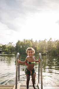 Smiling shirtless boy holding railing while standing on jetty near lake during vacation