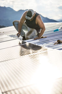 Worker uses hand tool to secure solar panels on roof of building.