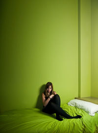 Portrait of young woman sitting on bed against wall