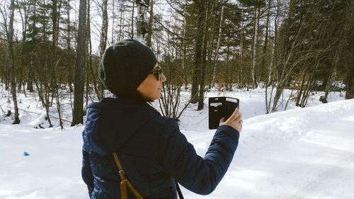 Rear view of woman with smart phone standing at forest during winter