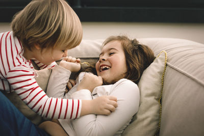 Playful girl tickling cheerful sister on couch at home