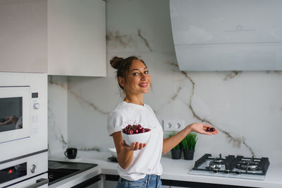 Pretty smiling young woman holds a plate of fresh cherry berries in her hand in the kitchen