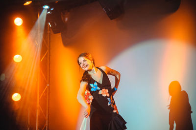 Portrait of smiling young woman standing against illuminated stage