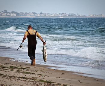 Rear view of man holding fish as he walks on beach