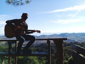 Man playing guitar while sitting on railing against sky