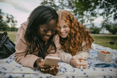 Happy teenage girl lying beside female friend using smart phone on picnic blanket