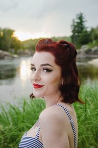 Portrait of beautiful woman standing on grassy field by lake against sky