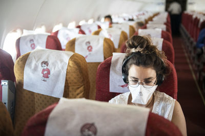 Young woman wearing a face mask while she listens to music on an almost empty plane, consequence of the coronavirus pandemic of 2020.