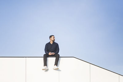 Contemplating man looking away while sitting on built structure against blue sky