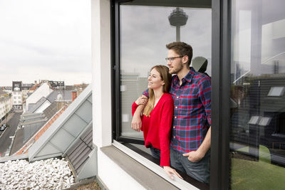 Smiling young couple looking out of window in city apartment