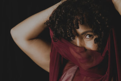 Portrait of beautiful woman with thin cloth of dark red color on her face against black background. 