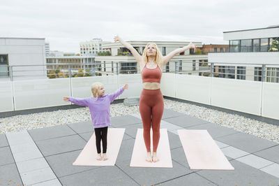 Daughter with arms outstretched looking at mother practicing yoga on rooftop