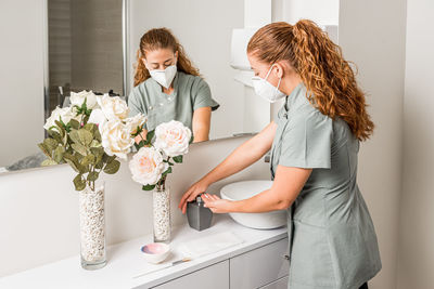 Side view of young female beautician doctor in protective mask washing hands while preparing for treatment procedure in modern office