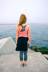 Rear view of young woman standing on retaining wall against sea