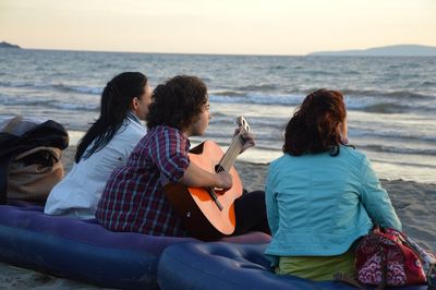 Rear view of friends with guitar sitting at beach against sky