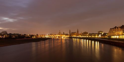 Illuminated buildings by river against sky at sunset