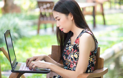 Side view of young woman using laptop while sitting on chair in yard