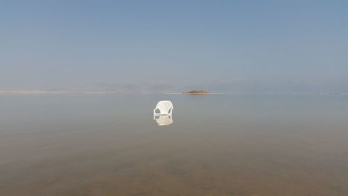 Empty abandoned white chair in sea against clear sky during sunny day