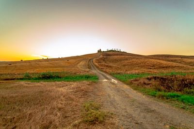 Dirt road amidst field against sky during sunset