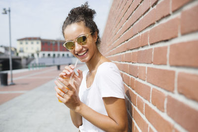 Happy young woman leaning against a brick wall holding orange juice