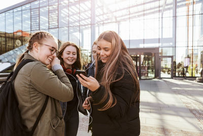 Teenage girls listening music through mobile phone while standing with friends outside railroad station