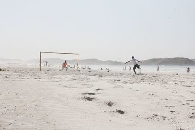 Male friends playing soccer at beach against clear sky