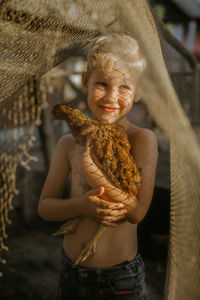 Smiling shirtless boy holding chicken while standing outdoors