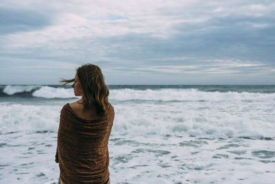 Rear view of mid adult woman with scarf standing at beach against sky