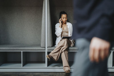 Female entrepreneur talking on smart phone while sitting with legs crossed in booth at office