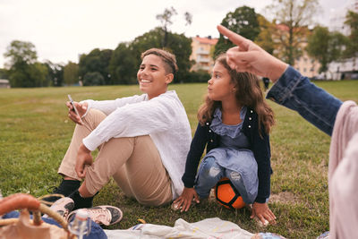 Siblings looking away while sitting on grassy field in park during picnic