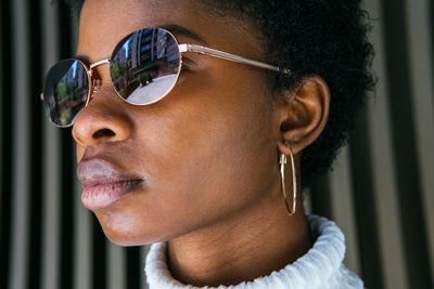 Side view of young african american woman in stylish sweater and sunglasses looking away while standing in bright sunlight against black background