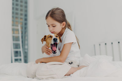 Cute girl embracing dog while sitting on bed at home