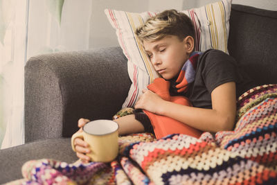 Sick boy holding milk cup while lying don on sofa at home