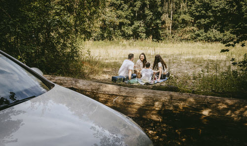 People sitting by car on road against trees