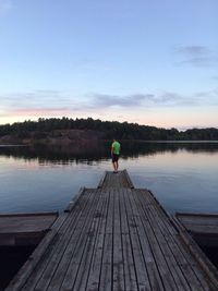 Full length rear view of man standing on pier by lake against sky at dusk