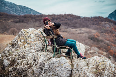 Man photographing on mobile phone while sitting on rock against mountain