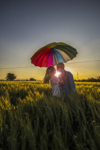 Romantic couple holding umbrella standing on field against sky during sunset